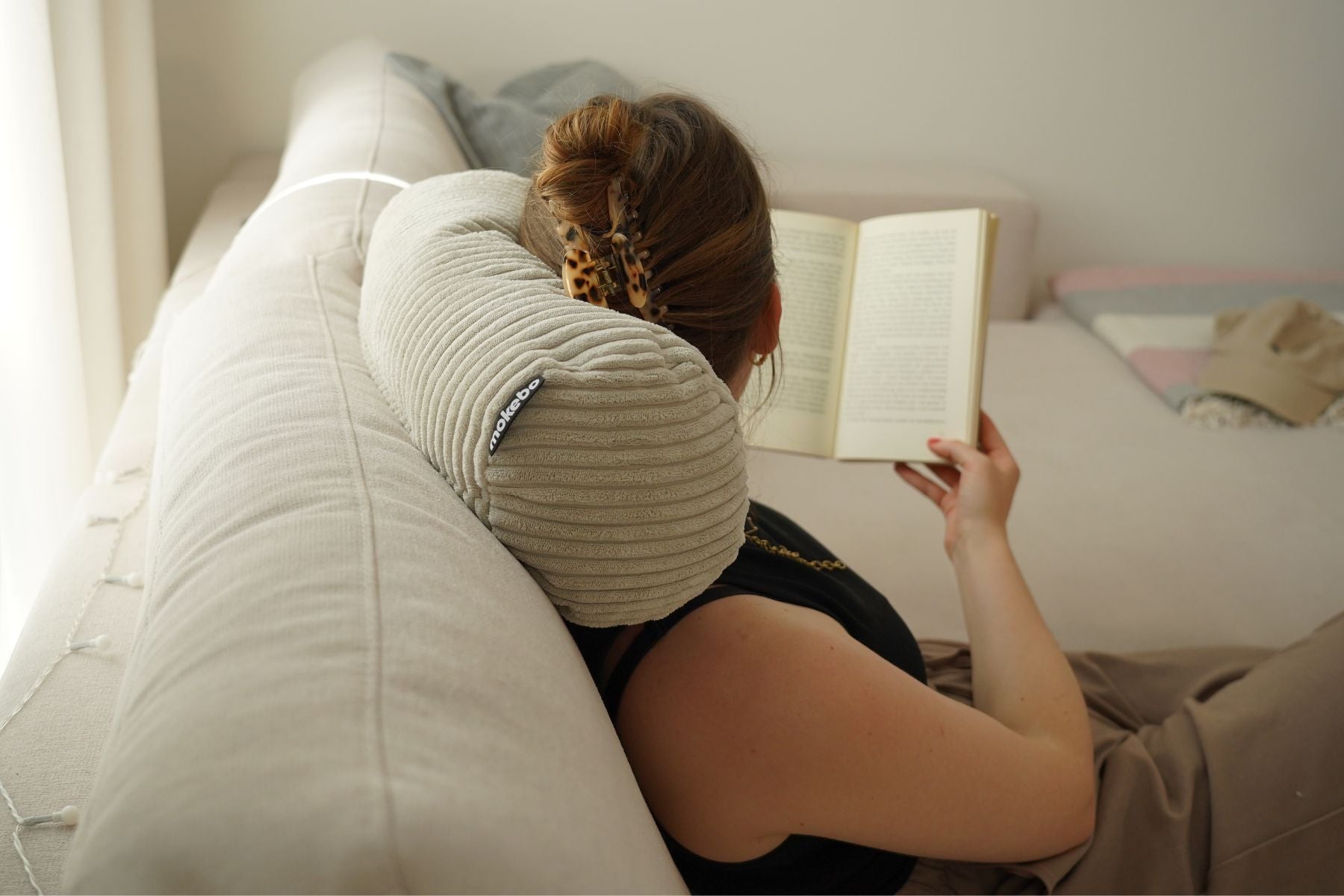 Nackenkissen in Cord beige. Kissen liegt im Nacken von Frau. Frau liest und schaut von der Kamera weg. 